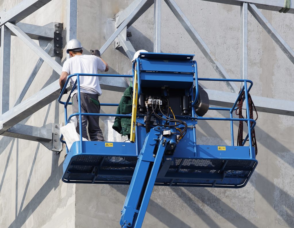 A worker wearing a helmet stands on a blue hydraulic lift, inspecting or working on a metal structure attached to a concrete wall.
