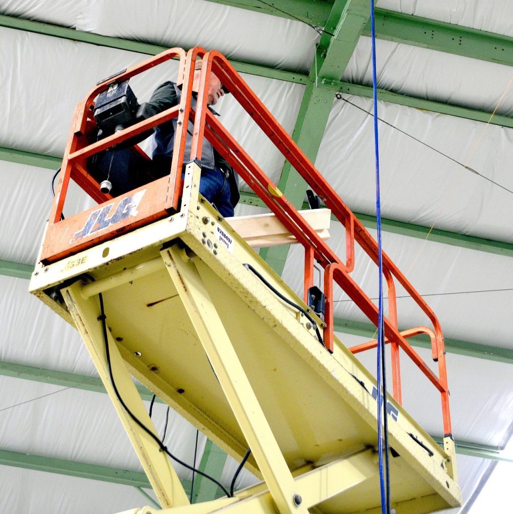 A person operates a scissor lift inside a large building with a green metal frame and insulated ceiling.
