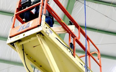 A person operates a scissor lift inside a large building with a green metal frame and insulated ceiling.